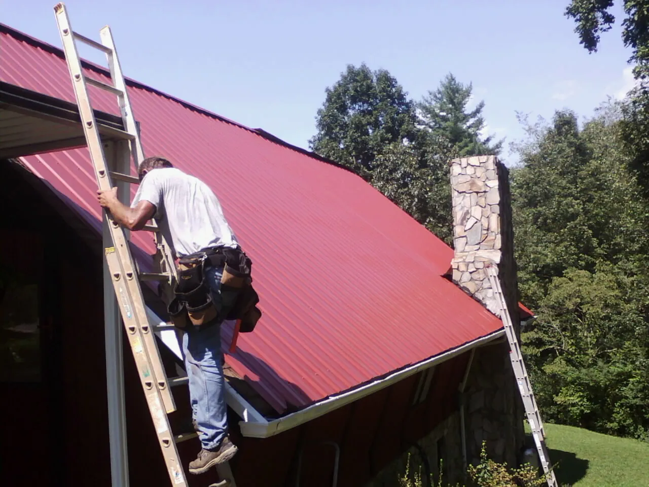 Roofer on a ladder beside a red metal roof and stone chimney — Soffit & Fascia Repair by Roofing Brothers in Mico